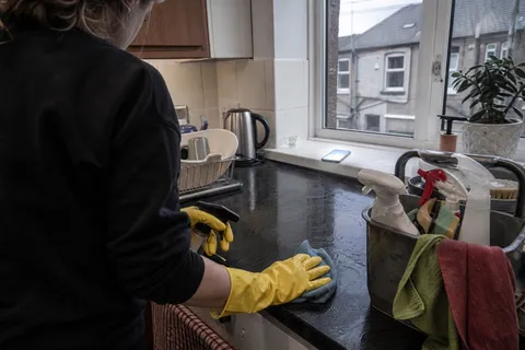 A cleaner in rubber gloves wiping a kitchen worktop while a mobile phone lights up and buzzes unanswered on the windowsill, the moment a cleaning business misses an enquiry call.