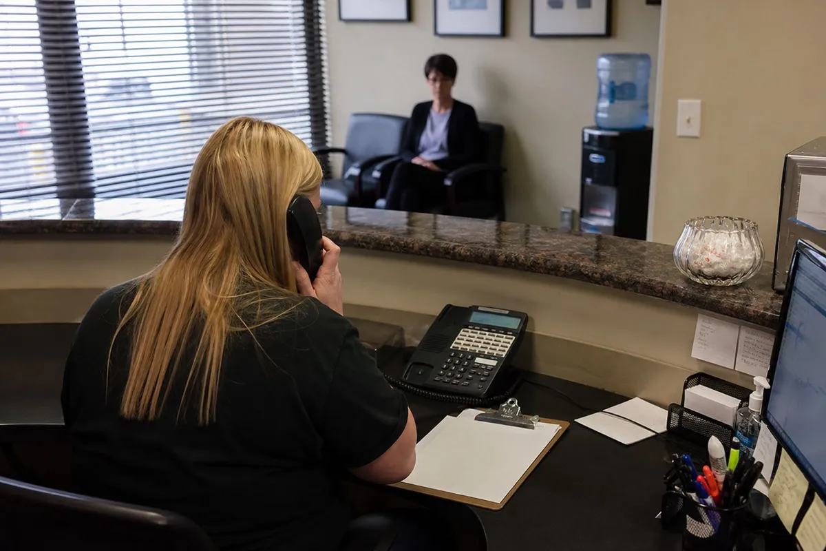 A dental receptionist answering the phone at a busy practice front desk while a patient waits.