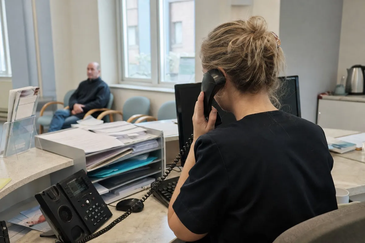 A dental receptionist answering the phone at a busy practice front desk while a patient waits.