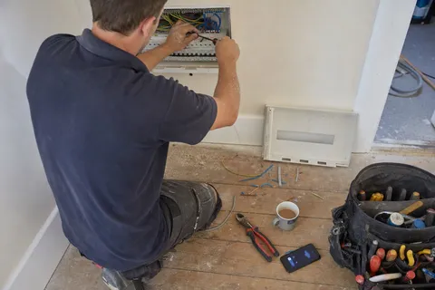 An electrician crouched at a domestic consumer unit with both hands on the wiring while a mobile phone lights up unanswered on the floor beside the toolbag