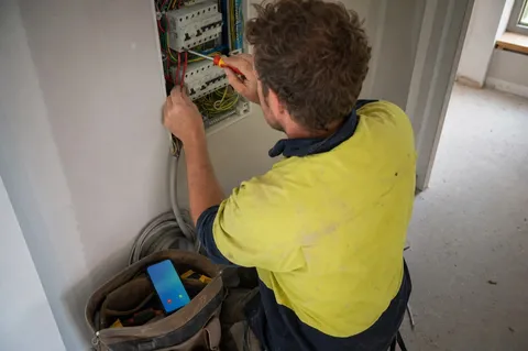 An electrician kneeling at an open domestic consumer unit with both hands busy, while a phone lights up unanswered on the toolbag beside him.