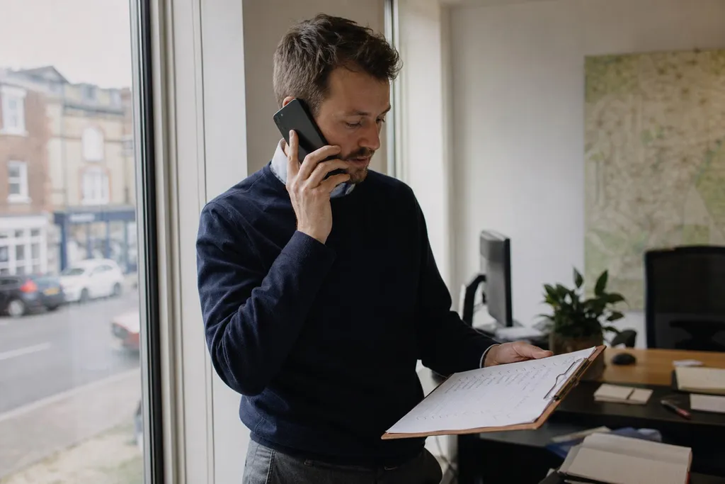 An estate agent making a follow-up call by the office window with a list of contacts on a clipboard
