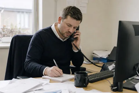 An estate agent at their desk taking a buyer enquiry on the phone and jotting down notes