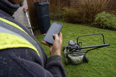 A landscaper pauses mowing to glance at a missed call on a mud-flecked phone pulled from a work trouser pocket