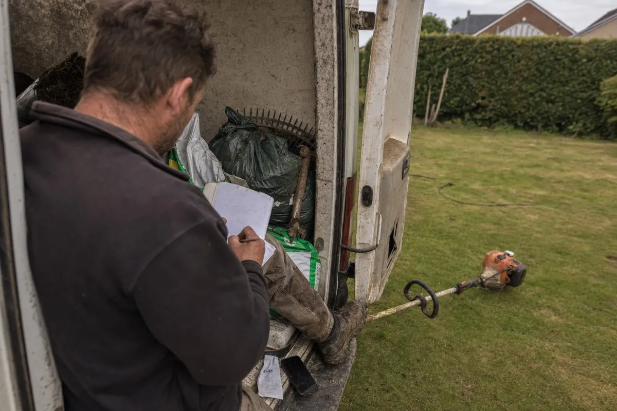 A landscaper sitting on the back of a work van, writing job costs and a quote on a paper notepad