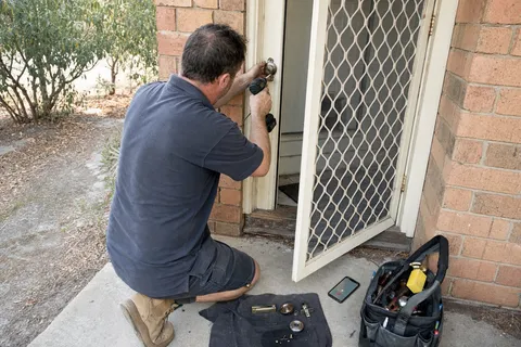 A one-van locksmith kneeling at a front door working on the lock while his phone rings unanswered on the step beside his tools
