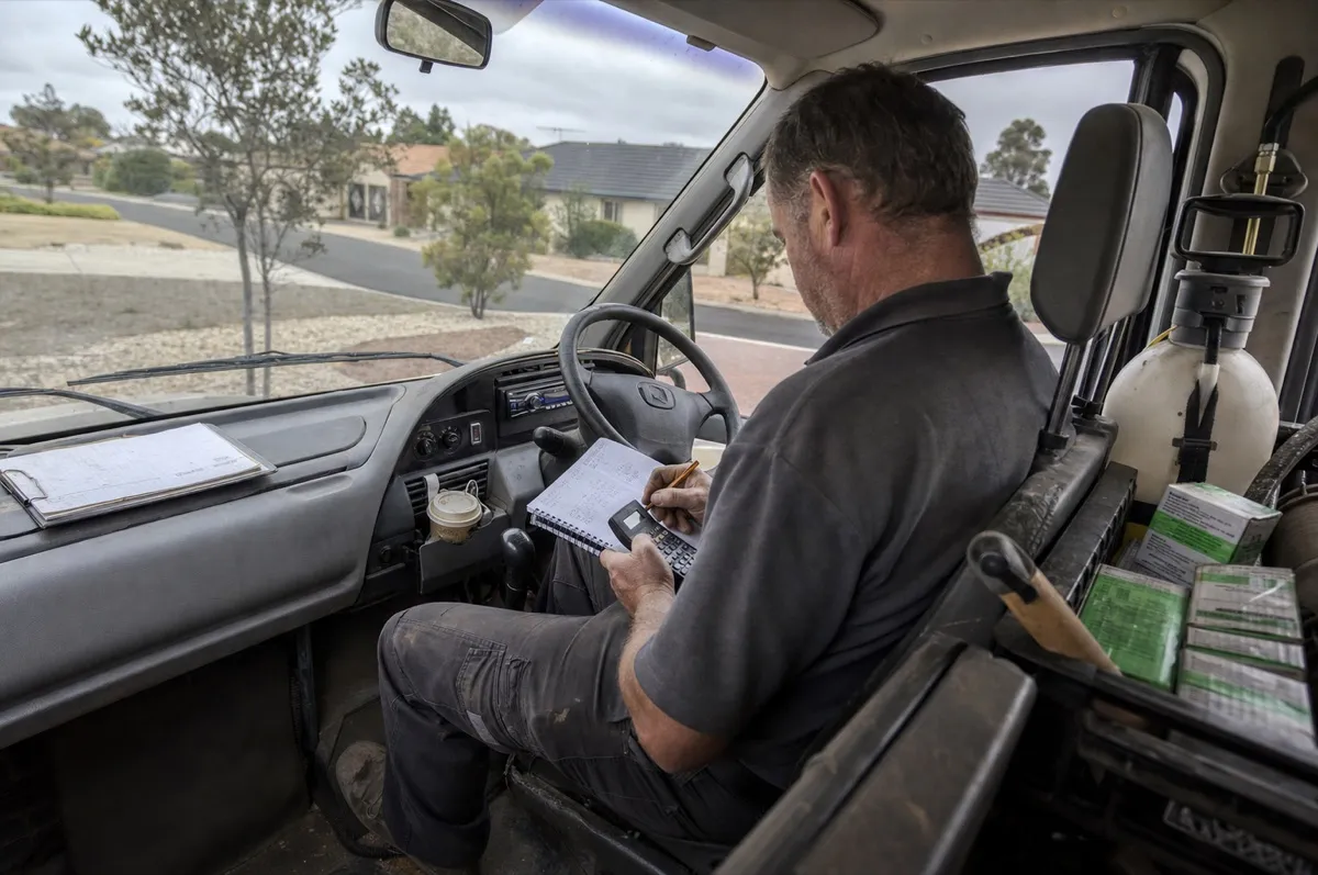 A pest control technician sitting in his work van, writing figures in a notebook with a calculator to work out the price for a job.