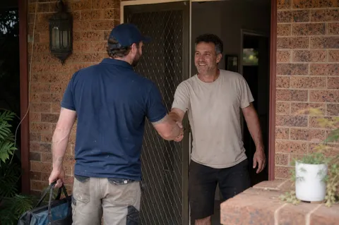 A plumber greeting a friendly homeowner at the front door of an Australian home, toolbag in hand