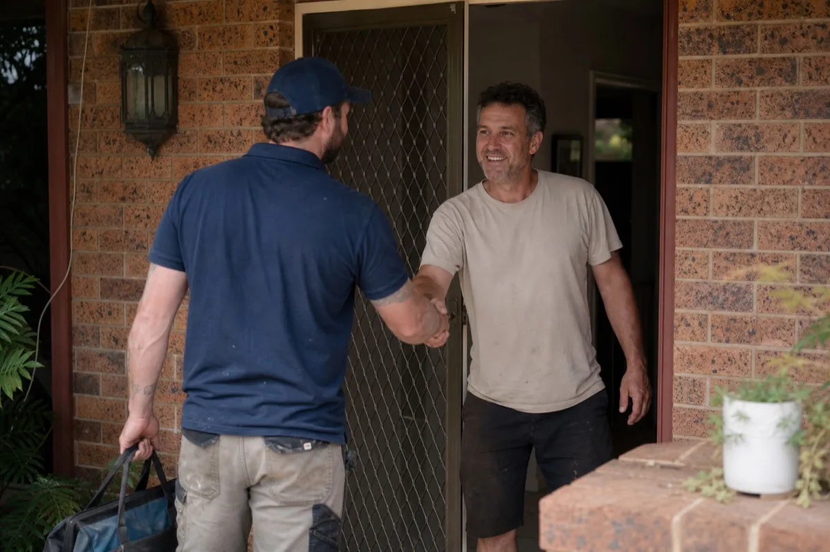 A plumber greeting a friendly homeowner at the front door of a New Zealand home, toolbag in hand