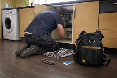 A plumber working under a kitchen sink while a phone rings unanswered on the floor beside the toolbag