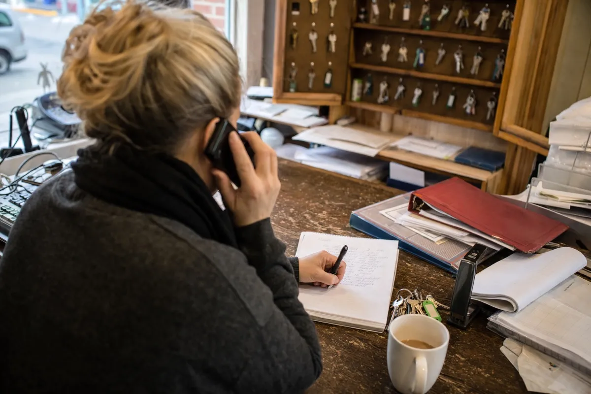 A property manager holds a phone to their ear and writes a repair note on a pad at a desk, with a board of labelled property keys behind them.