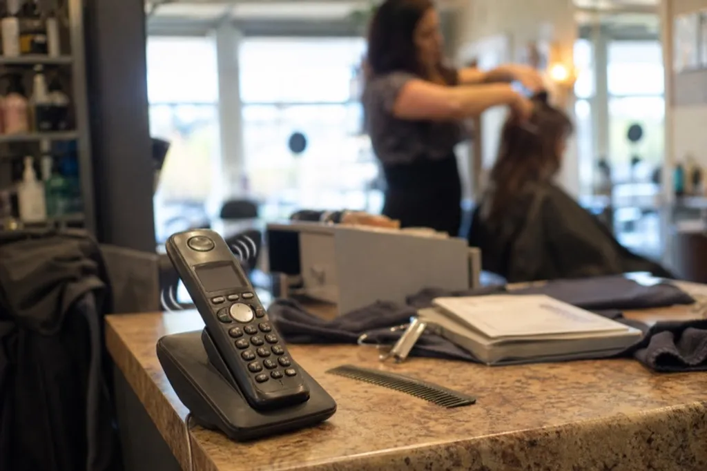 A salon reception phone ringing unanswered while a stylist works at the chair behind it