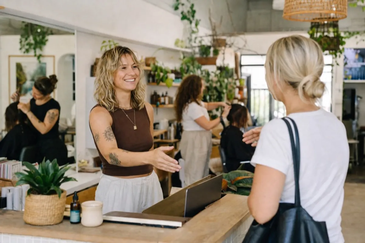 A salon stylist welcoming a new client at the front desk of a busy New Zealand hair salon