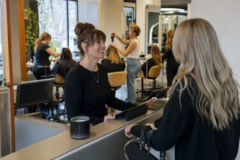 A salon stylist welcoming a new client at the front desk of a busy American hair salon