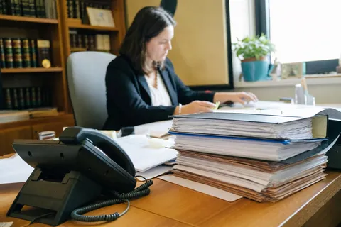 A solicitor at a desk stacked with case files and ring binders next to a desk phone in a small law office.