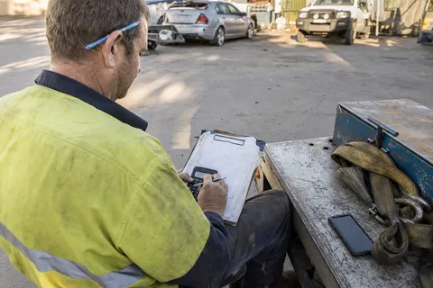 A recovery truck operator sits on the flatbed of his tow truck working out the day's takings on a clipboard with a calculator.