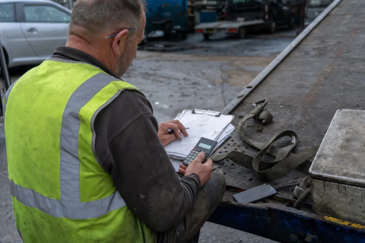 A recovery truck operator sits on the flatbed of his tow truck working out the day's takings on a clipboard with a calculator.
