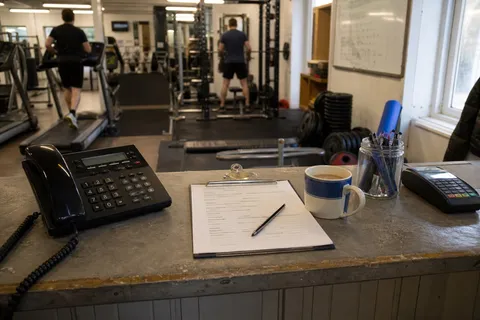 An unattended front desk in a small gym with a phone on the counter and members training in the background